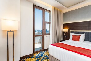 Une chambre d’hôtel avec un grand lit, une vue sur la ville par la fenêtre, une lampe sur pied et un tapis aux motifs bleus et jaunes.