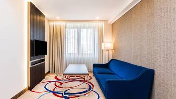 Quarto de hotel com televisão de tela plana fixada em painel de madeira, sofá azul, mesinha e luminária de chão.