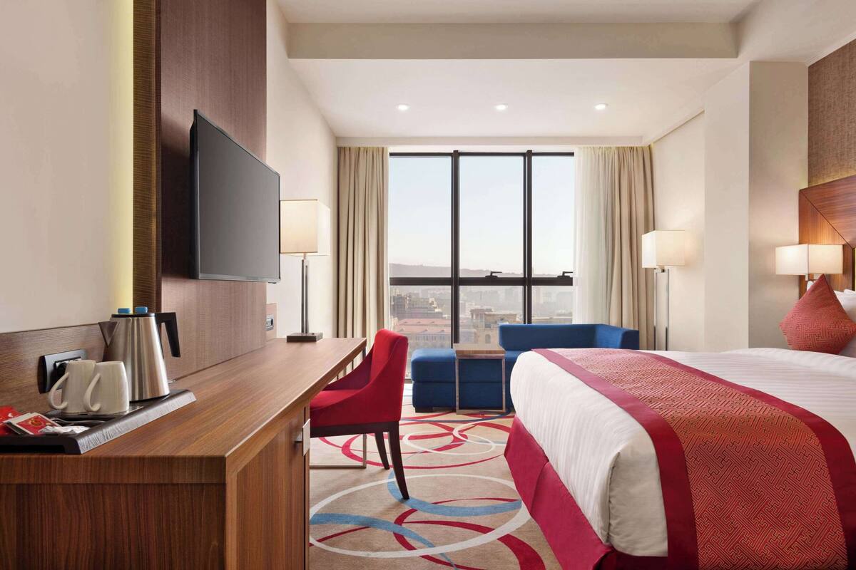 Une chambre d’hôtel avec un grand lit, un bureau avec une cafetière, une télévision et une vue sur la ville grâce à de grandes fenêtres.