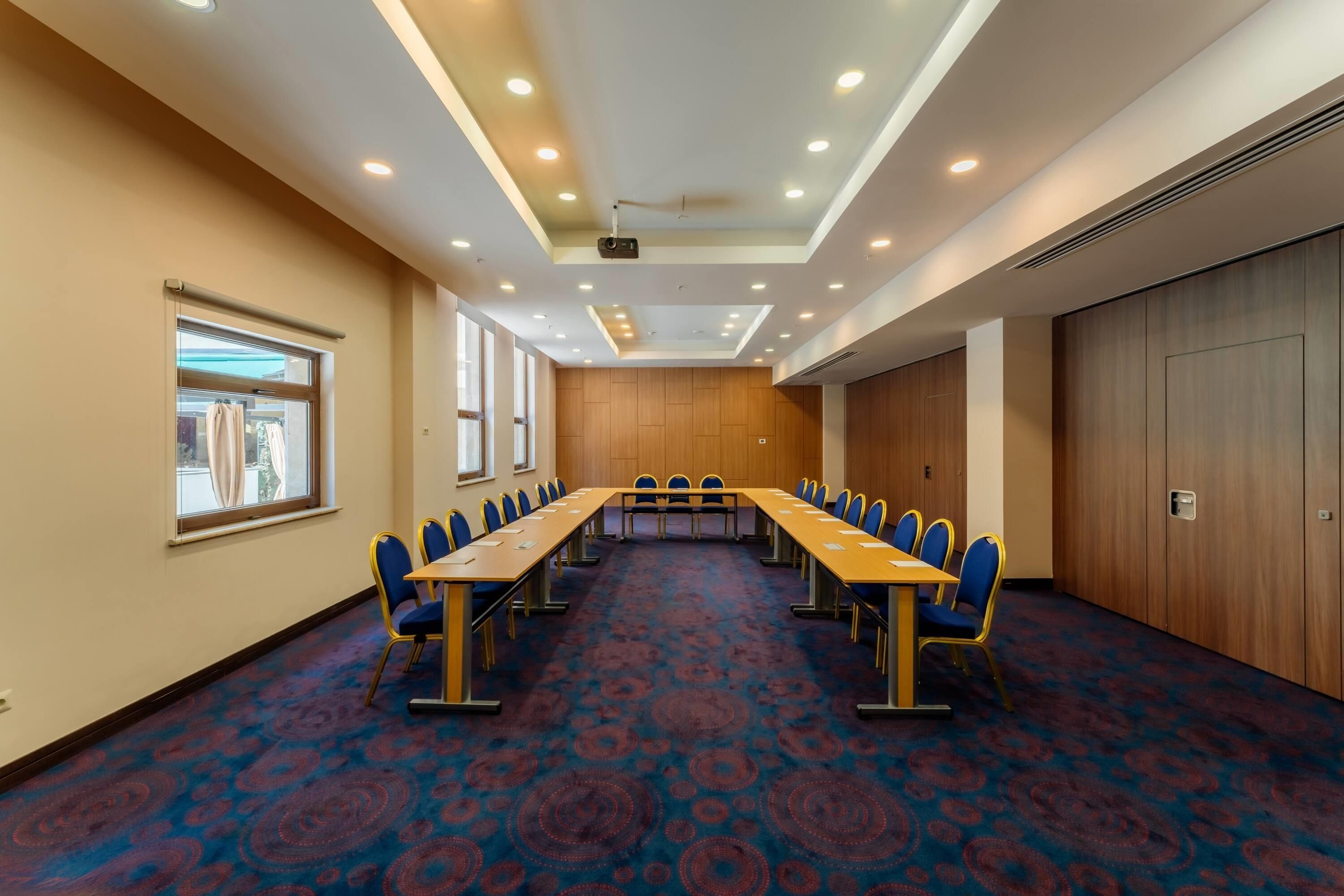A conference room with a long table and chairs, large windows, and a patterned carpet.