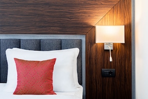 Une chambre d’hôtel avec un lit, un oreiller rouge à motifs, une tête de lit grise et une lampe fixée au mur.