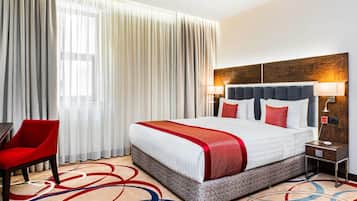 A hotel room with a large bed, a red chair, a wooden headboard, a nightstand, and a red and white patterned rug.