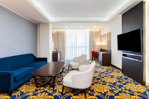 Een hotelkamer met een blauwe bank, een eettafel, twee fauteuils, een televisie en een groot raam met gordijnen.