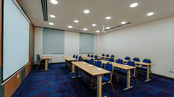 Een vergaderzaal met rijen tafels en stoelen, een scherm voor een beamer en een groot raam.