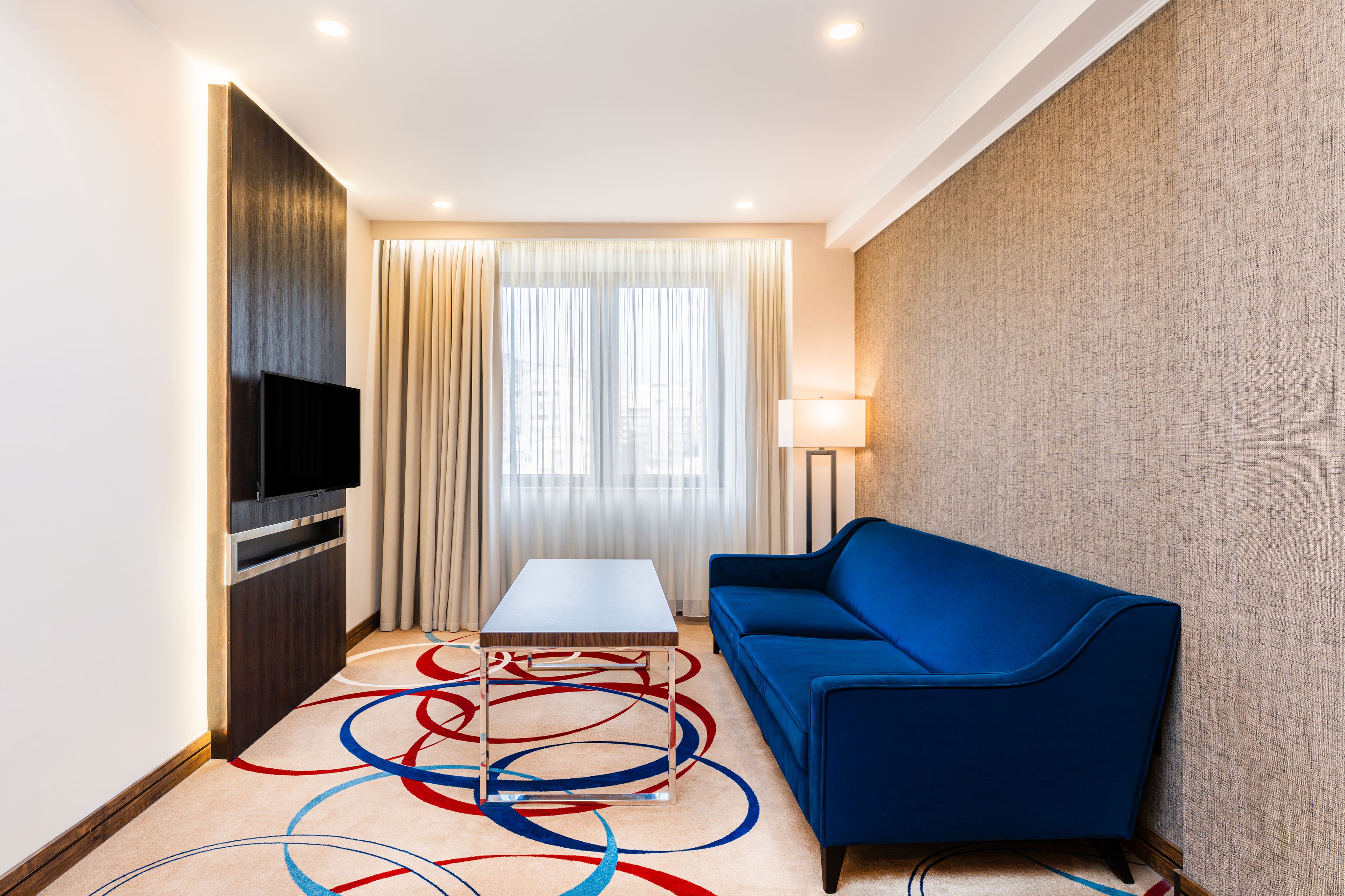 A hotel room with a flat-screen TV, a blue sofa, a small table, and a floor lamp.