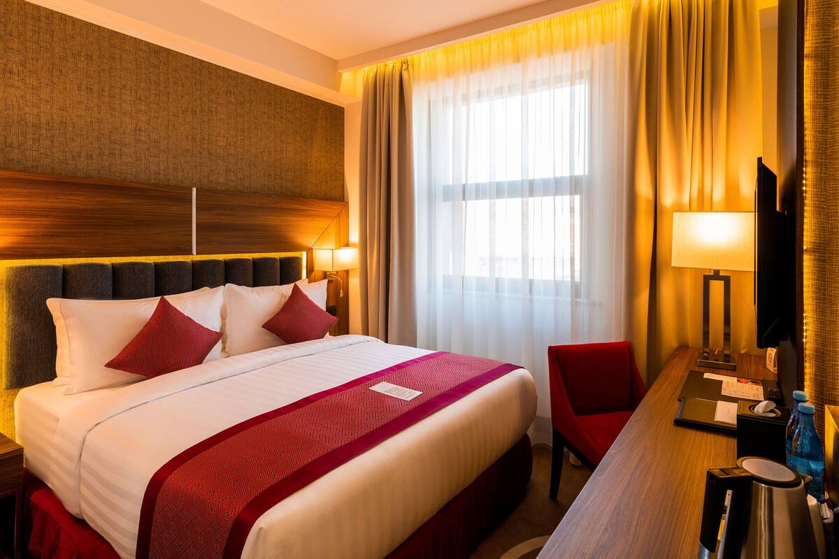 Une chambre d’hôtel avec un grand lit, une literie rouge et blanche, une tête de lit en bois, un bureau avec une chaise, une télévision et une fenêtre avec des rideaux.