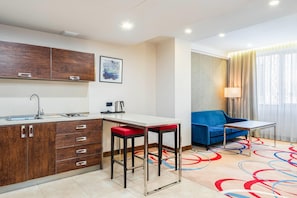 Une petite cuisine moderne équipée d’un évier, d’une cuisinière et de meubles en bois. On y trouve une petite table avec une chaise rouge, un canapé bleu et une lampe près d’une fenêtre ornée de rideaux.