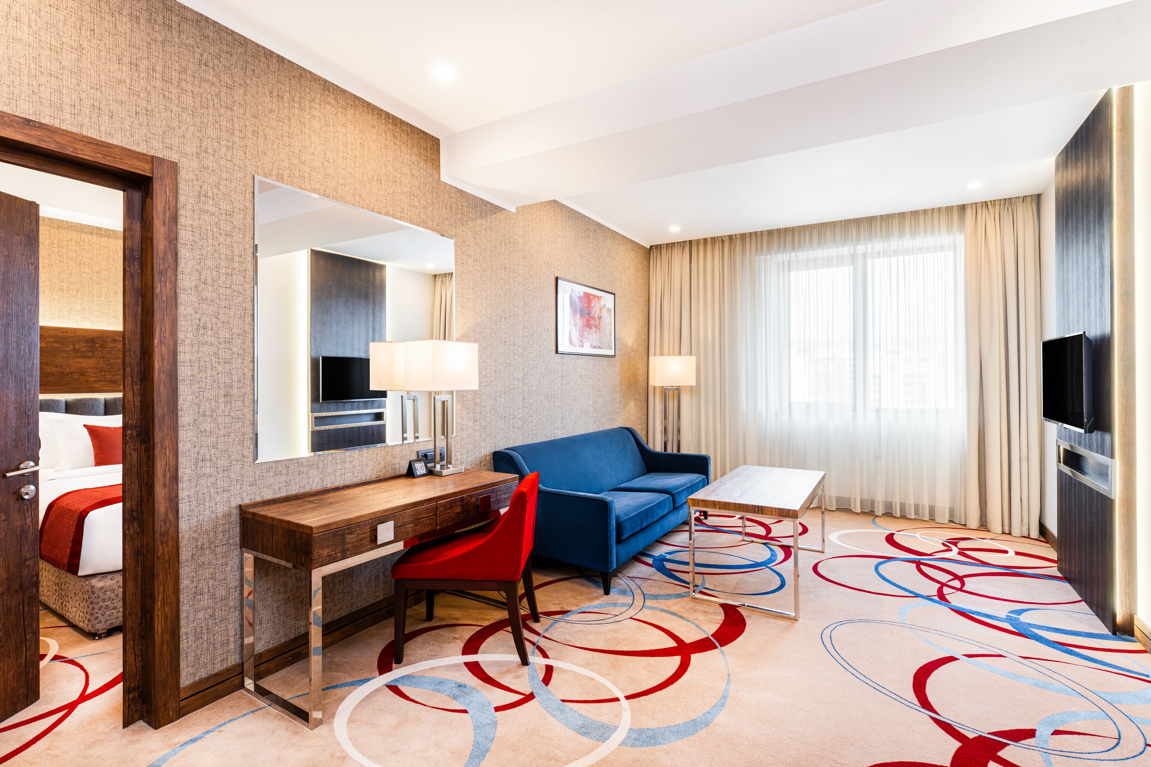 A hotel room with a wooden desk, a blue sofa, a small table, a TV, and a large window with curtains.