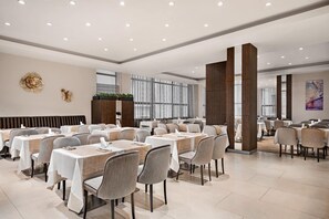 Un espace de restauration moderne, doté de plusieurs tables et chaises, d’un élément décoratif mural et de grandes fenêtres laissant entrer la lumière naturelle.