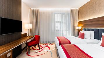 A hotel room with a flat-screen TV, a desk with a chair, a bed with red and white bedding, and a large window with sheer curtains.
