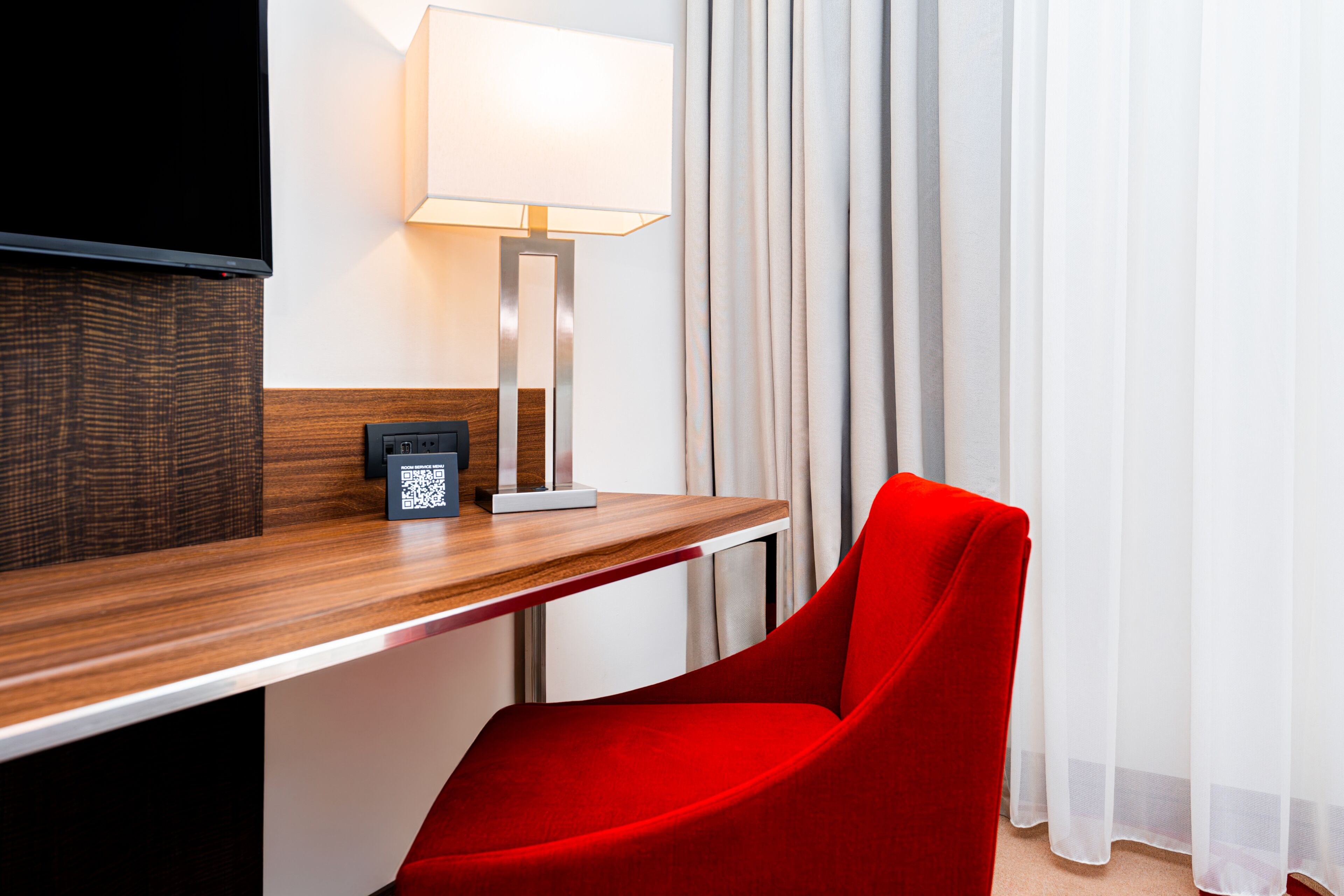 Habitación de hotel con un escritorio de madera, una silla roja, una lámpara, un televisor de pantalla plana y una ventana con cortinas.