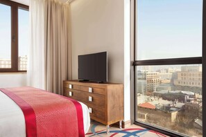 Ett hotellrum med en säng, en byrå i trä, en tv och ett stort fönster med utsikt över staden.
