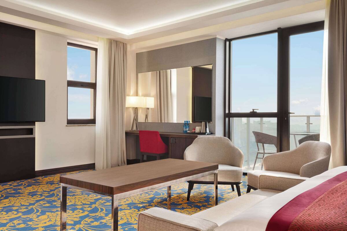 Une chambre d’hôtel dotée d’un grand lit, de deux fauteuils, d’un bureau avec une chaise, d’une télévision et de grandes fenêtres offrant une vue panoramique.