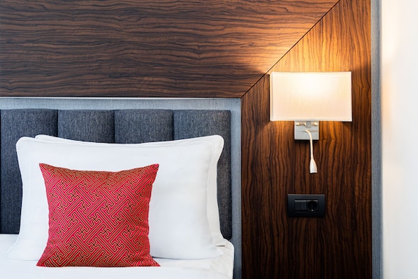 Habitación de hotel con cama, almohada roja estampada, cabecera gris y lámpara fijada a la pared.