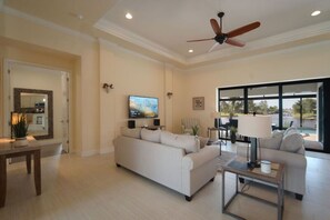 A spacious living room with a large sofa, a coffee table, and a TV. There is a ceiling fan, a large window with a view, and a door leading to another room.