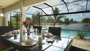 A dining area with a glass table and chairs, a vase with flowers, and a view of a water body and greenery. - Gulf access home, private pool, Jacuzzi, great waterview, free WIFI (Cape Coral,)