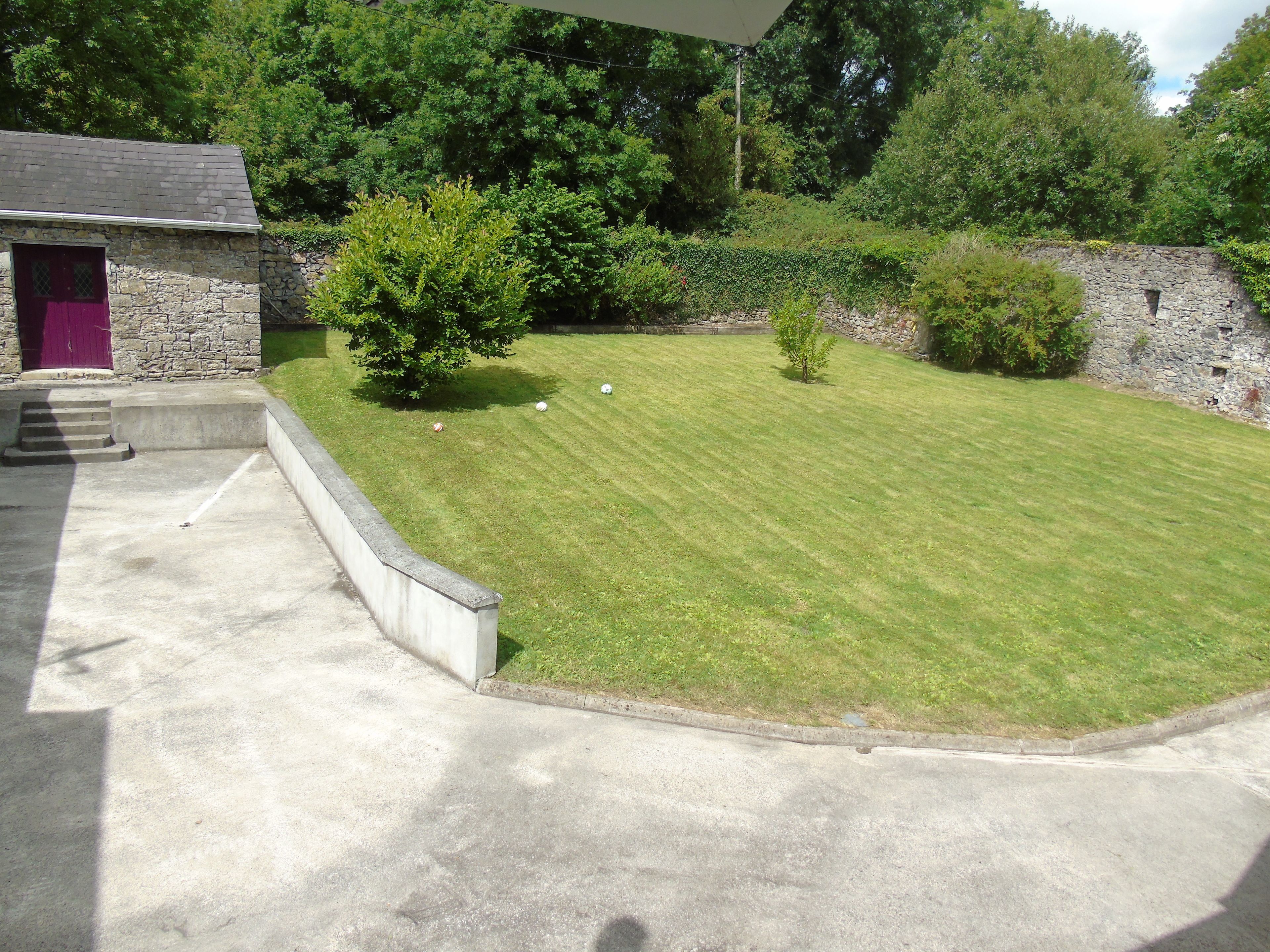 A stone building with a purple door, a small lawn with a few trees, and a stone wall in the background.