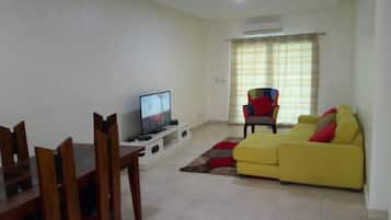 Uma sala de estar com um sofá amarelo, uma televisão em um móvel, uma mesa de jantar com cadeiras e uma poltrona colorida.