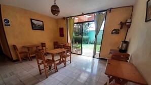Un espace repas avec des tables et des chaises en bois, un luminaire suspendu et une vue sur l’extérieur grâce à une porte vitrée.