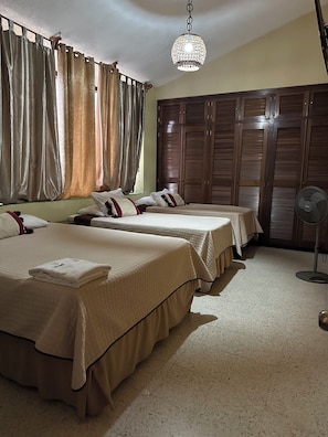 Une chambre d’hôtel avec deux lits, une grande armoire, un luminaire au plafond et un ventilateur sur pied.
