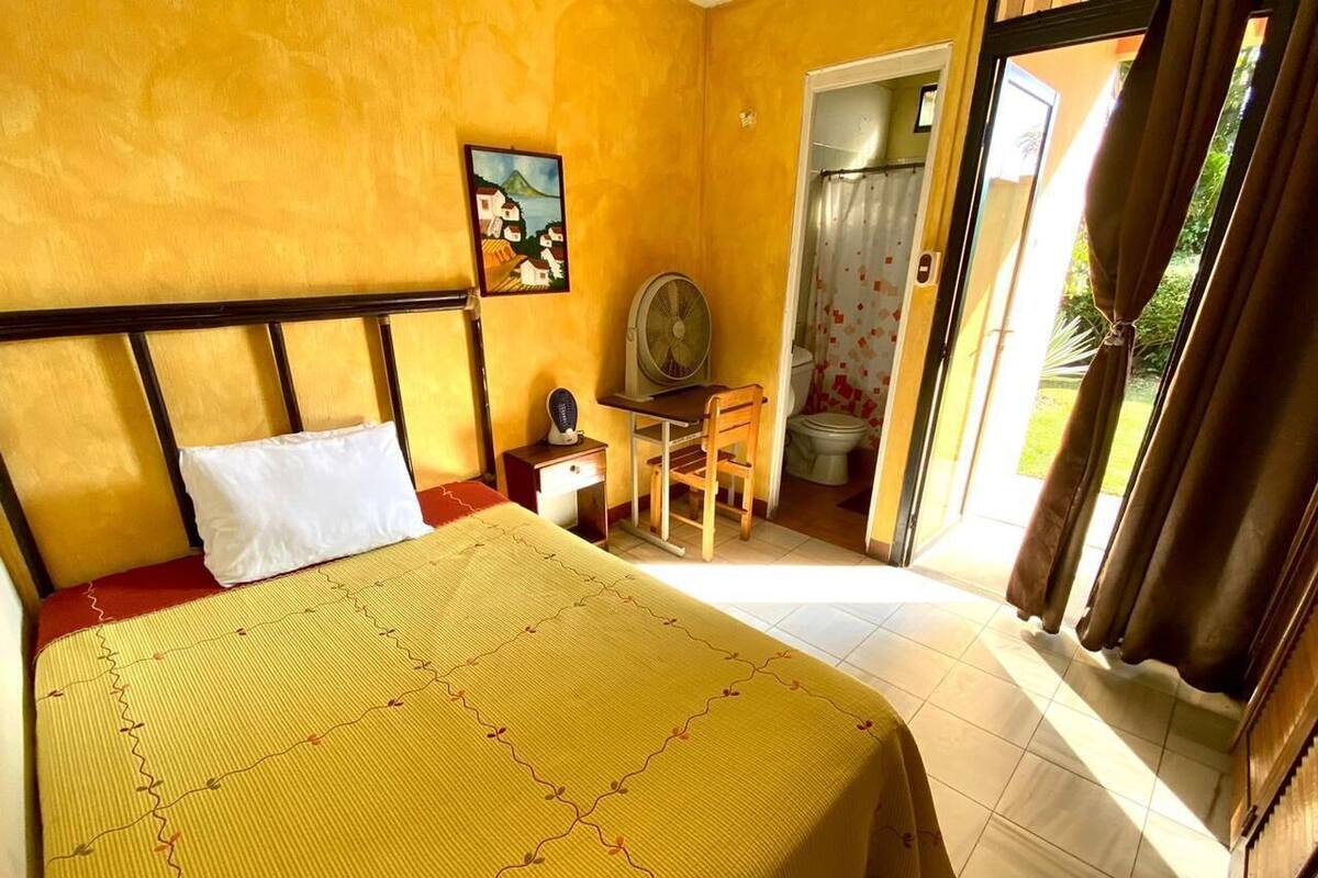 Ein Schlafzimmer mit Bett, Schreibtisch, Stuhl, Ventilator und einem durch eine offene Tür einsehbaren Badezimmer.