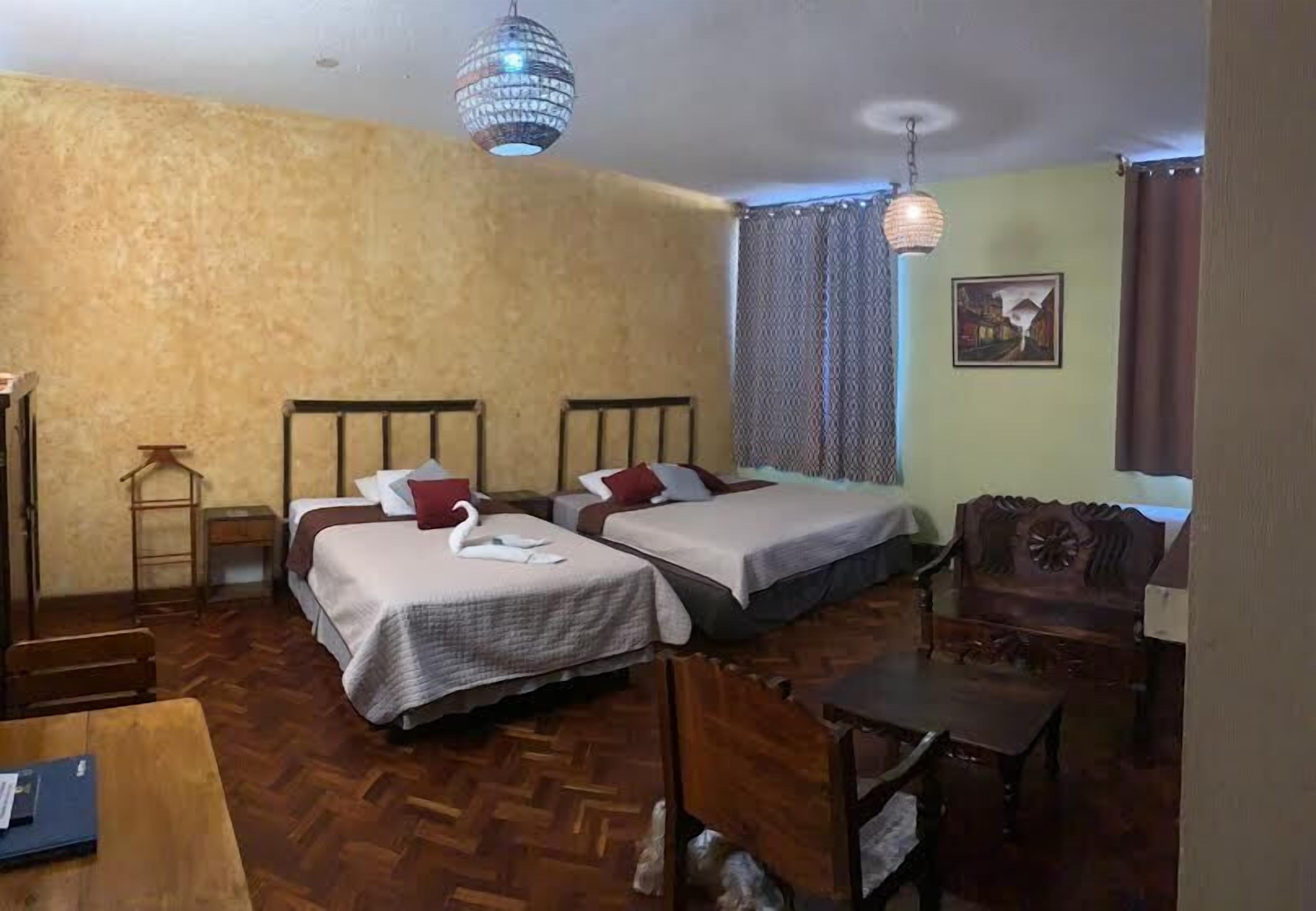 A room with two beds, a wooden table, a chair, a hanging light fixture, and a painting on the wall.