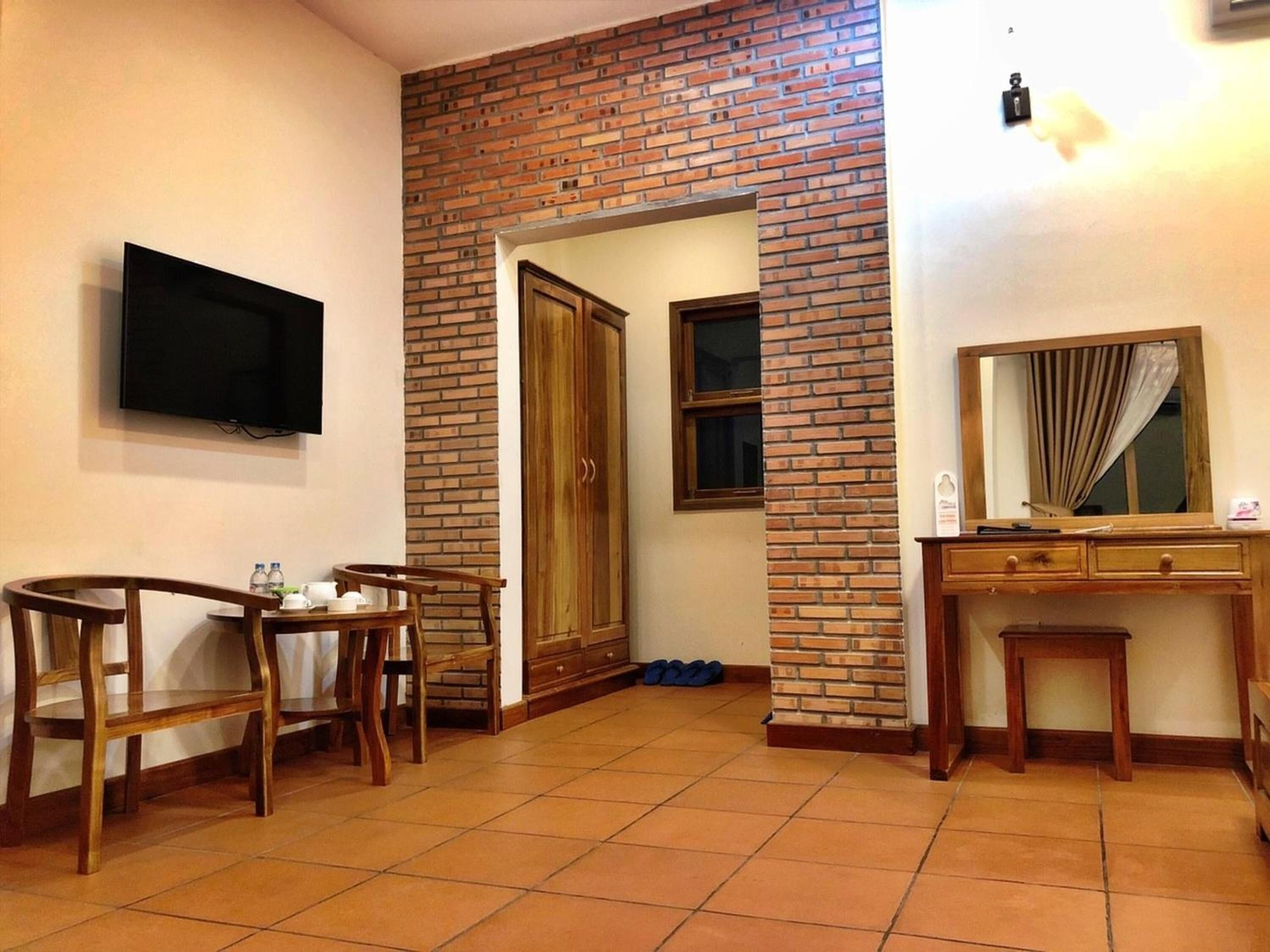 A room with a brick wall, a television mounted on the wall, a wooden desk with a mirror, and a wooden chair.