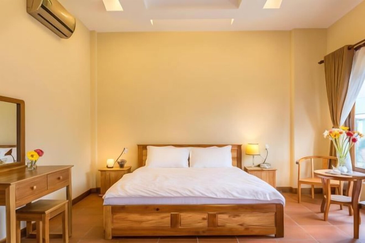 A hotel room with a wooden bed, a nightstand with a lamp, a desk with a mirror, and a chair.