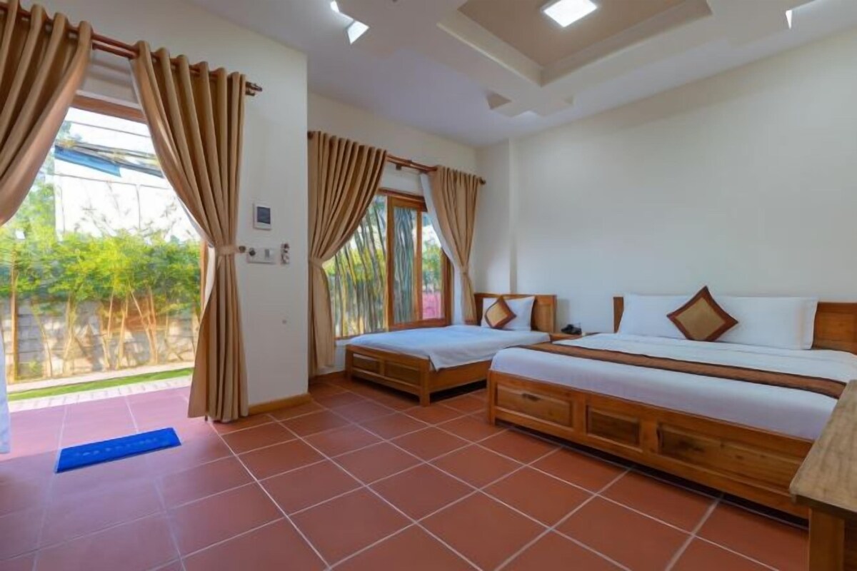A room with two beds, a window with curtains, a tiled floor, and a view of greenery outside.