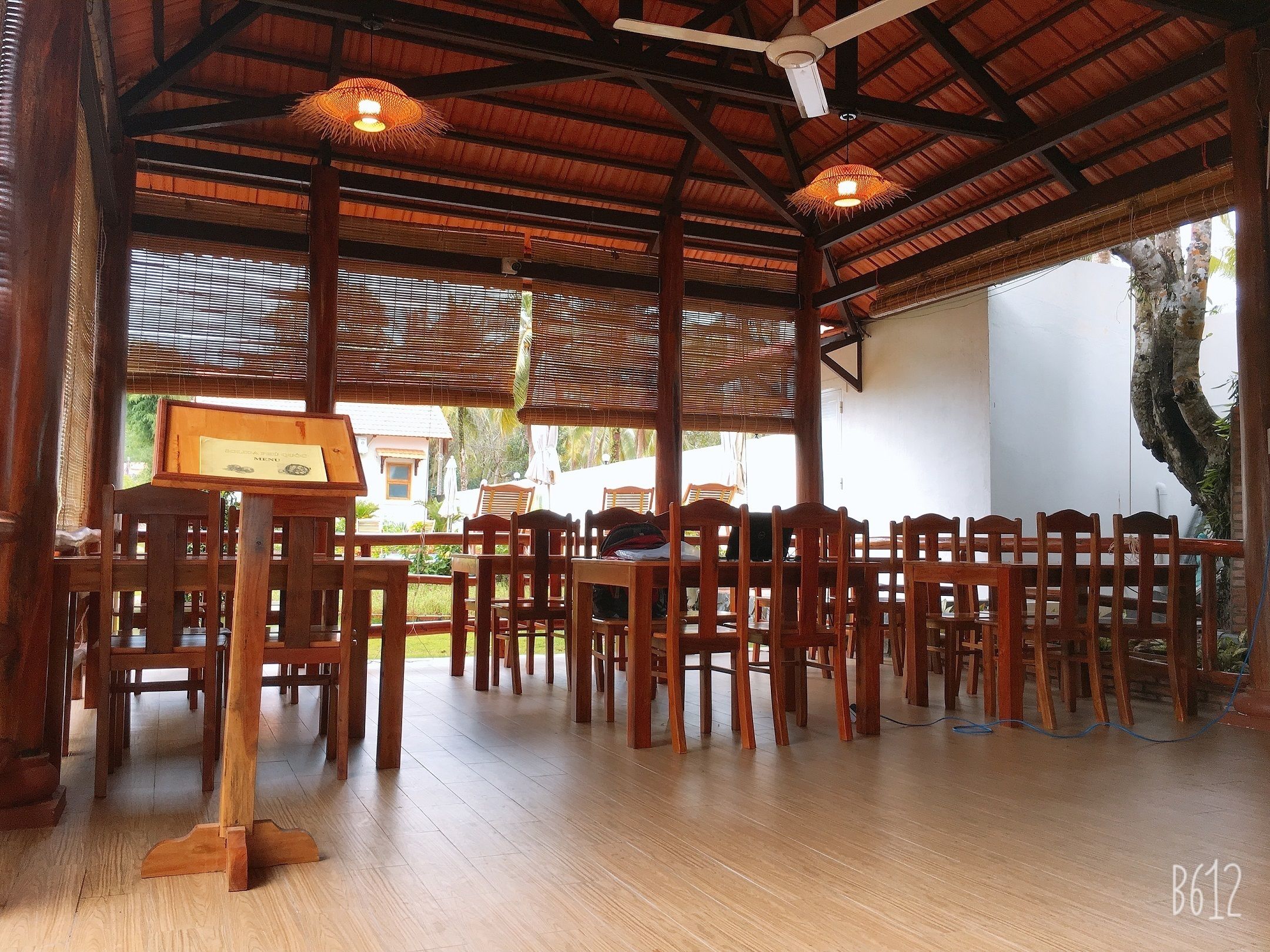 A covered outdoor dining area with wooden tables and chairs, a menu stand, and a view of the surrounding landscape.