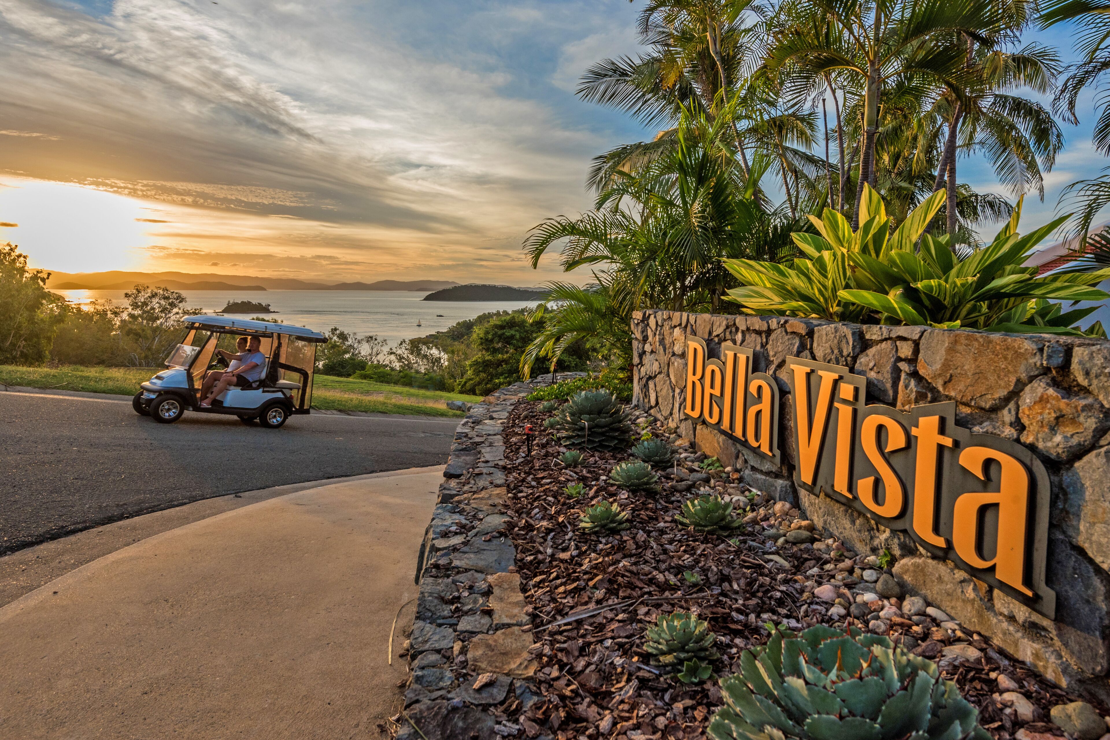 A golf cart parked near a sign reading "Bella Vista" with a stone wall and tropical plants.
