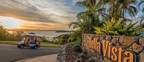 A golf cart parked near a sign reading "Bella Vista" with a stone wall and tropical plants.