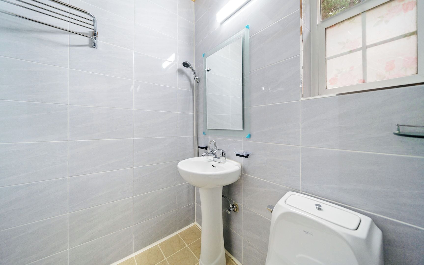 A bathroom with a pedestal sink, a handheld showerhead, a toilet, and a window with a floral view.