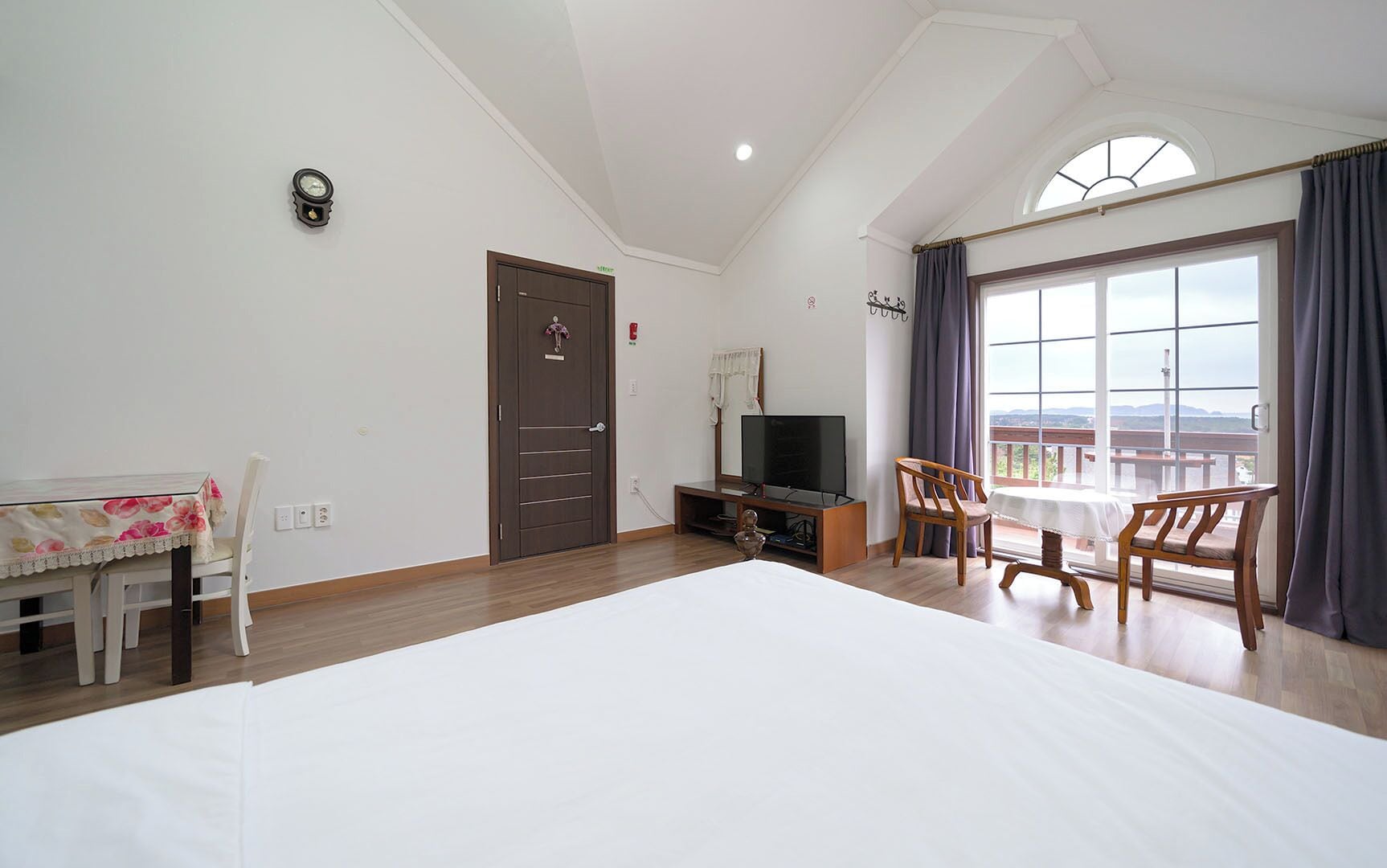 Un dormitorio con una cama grande, televisor, mesa de comedor con sillas, balcón con vistas y una puerta.