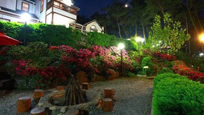 A well-lit garden with a fire pit, surrounded by stone benches and vibrant flowers. - Petit Maison Pension (Taean)