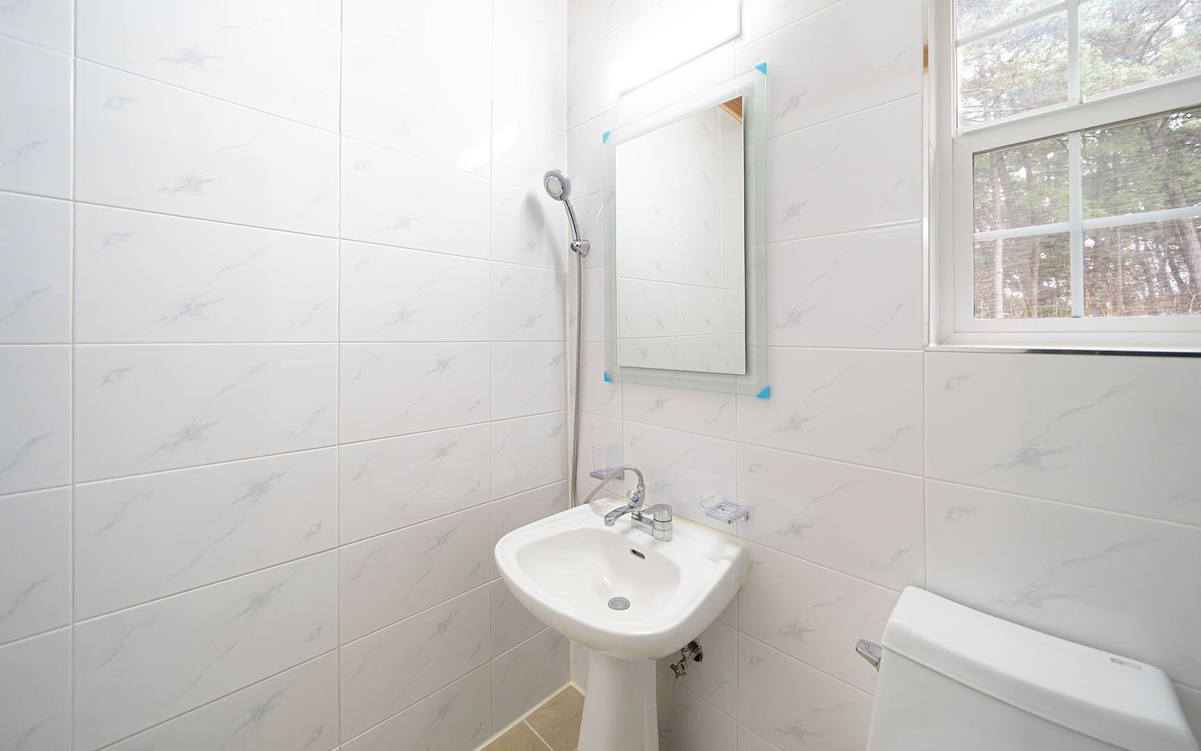 A bathroom with white marble tiles, a white sink, a mirror, and a window.