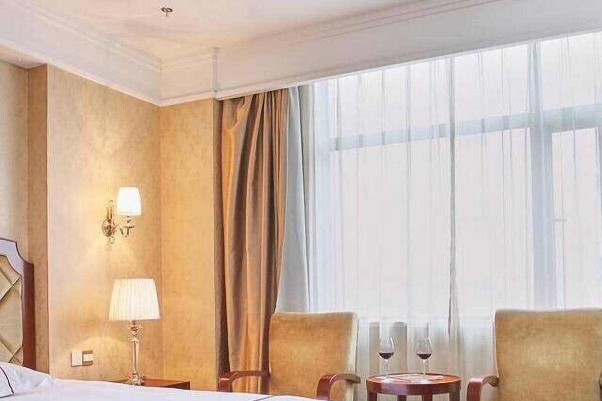 A hotel room with a large bed, two armchairs, a small table with a wine glass, and a window with sheer curtains.