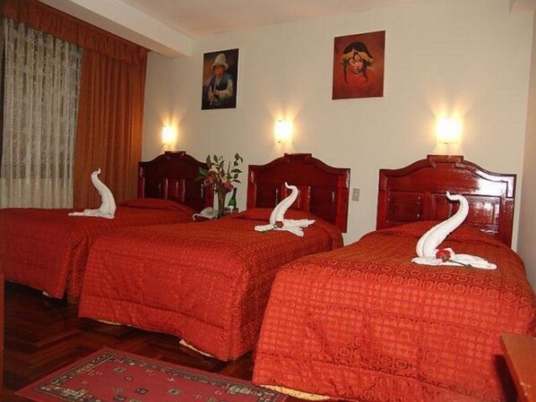 A hotel room with three beds, each with a red bedspread and a swan-shaped pillow. There are two wall-mounted lamps, a painting on the wall, and a wooden headboard. - inkayra hotel (Cusco)