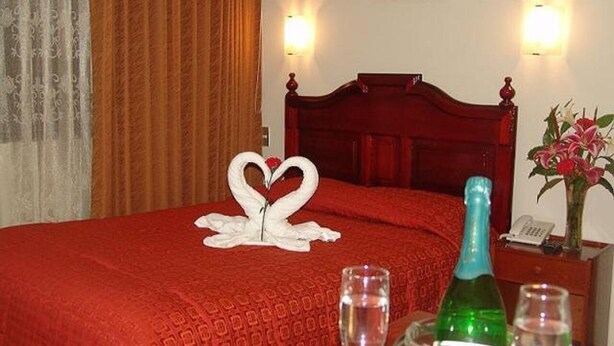 A hotel room with a red bedspread, a wooden headboard, two wall sconces, a bottle of champagne, and a vase with flowers. - inkayra hotel (Cusco)