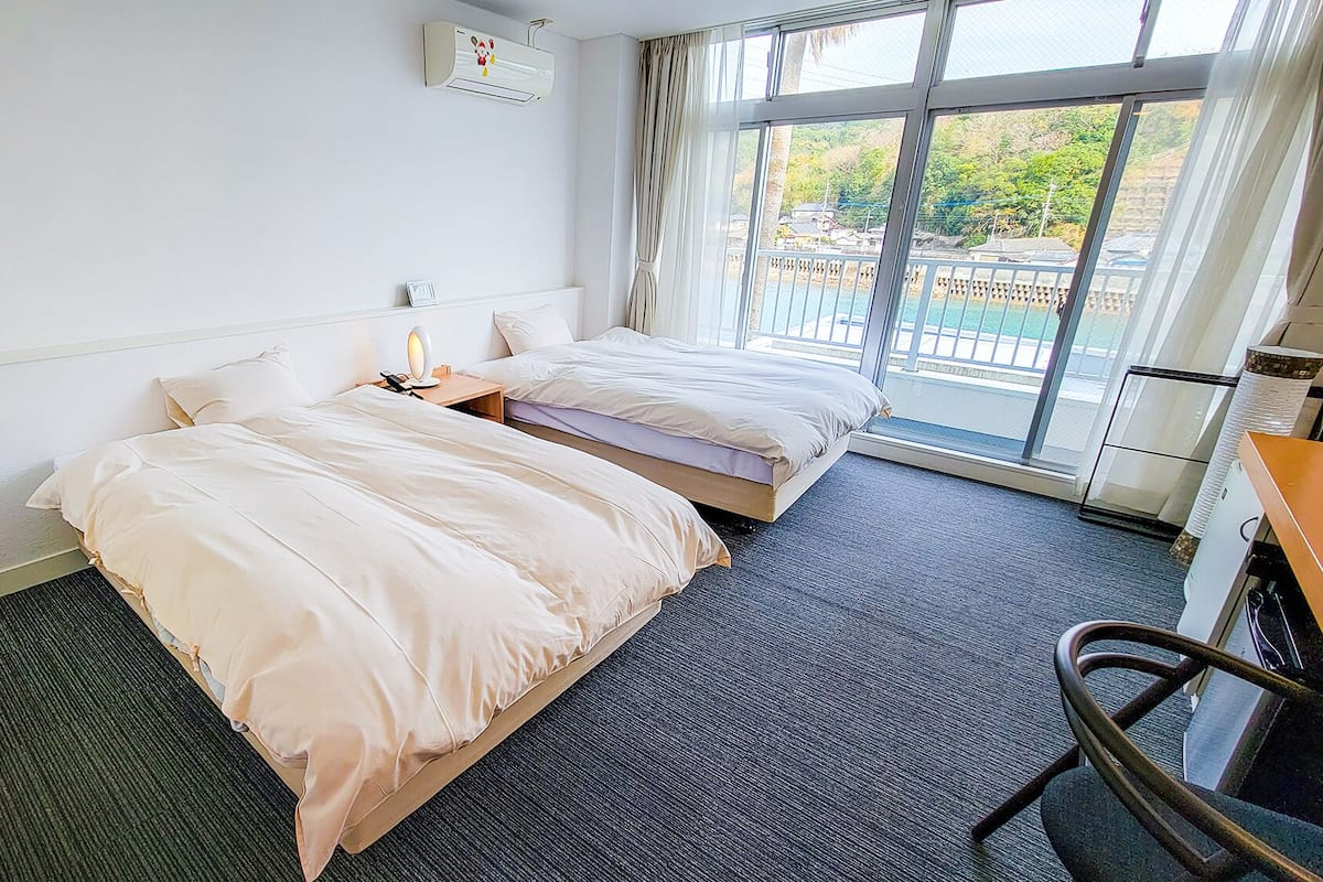 Une chambre d’hôtel avec deux lits, un bureau, une chaise, une fenêtre donnant sur un paysage agréable et un appareil de climatisation.