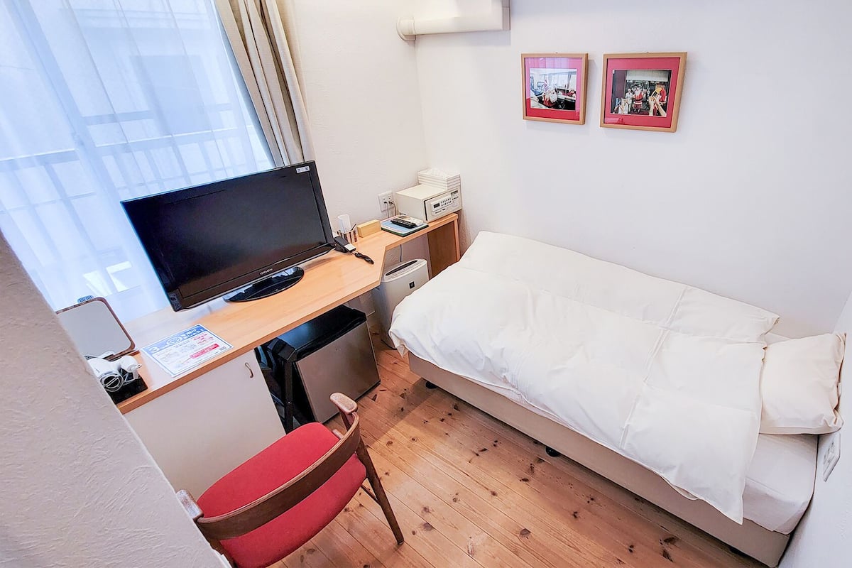 Une petite pièce, bien rangée, avec un lit, un bureau équipé d’un ordinateur, une chaise et deux tableaux encadrés accrochés au mur.