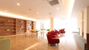 Um hall espaçoso com piso de azulejos brilhante, estantes de madeira com objetos decorativos, uma área de estar com cadeiras vermelhas e um balcão de recepção.