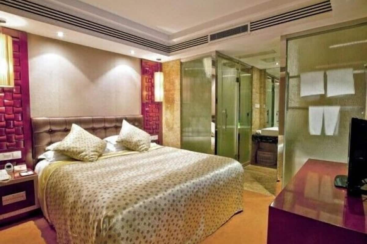 Quarto de hotel com uma cama grande, uma toucador com espelho, divisória de chuveiro em vidro e porta de acesso a um banheiro.