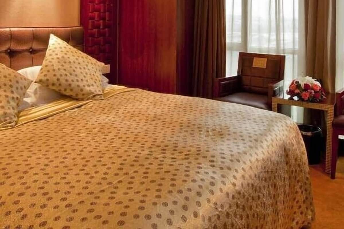 Quarto de hotel com uma cama grande, cabeceira de madeira, parede de destaque vermelha, janela com cortinas e uma mesinha com flores.