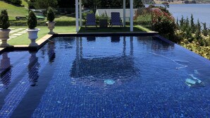 Una piscina infinita de color azul cristalino con un pabellón y dos sillas, rodeada de un jardín ajardinado y vistas al lago.