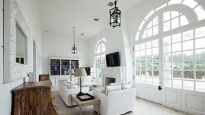 Amplia habitación con mobiliario blanco, chimenea y ventanas grandes que dan a zonas verdes.