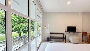 A hotel room with a bed, a TV, a small fridge, a bench, and large windows offering a view of greenery outside. - Be Leaf Resort (Thep Krasattri)