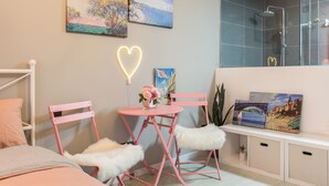 A neatly arranged bedroom with a bed, a pink chair, a small round table with a vase of flowers, and two paintings on the wall.