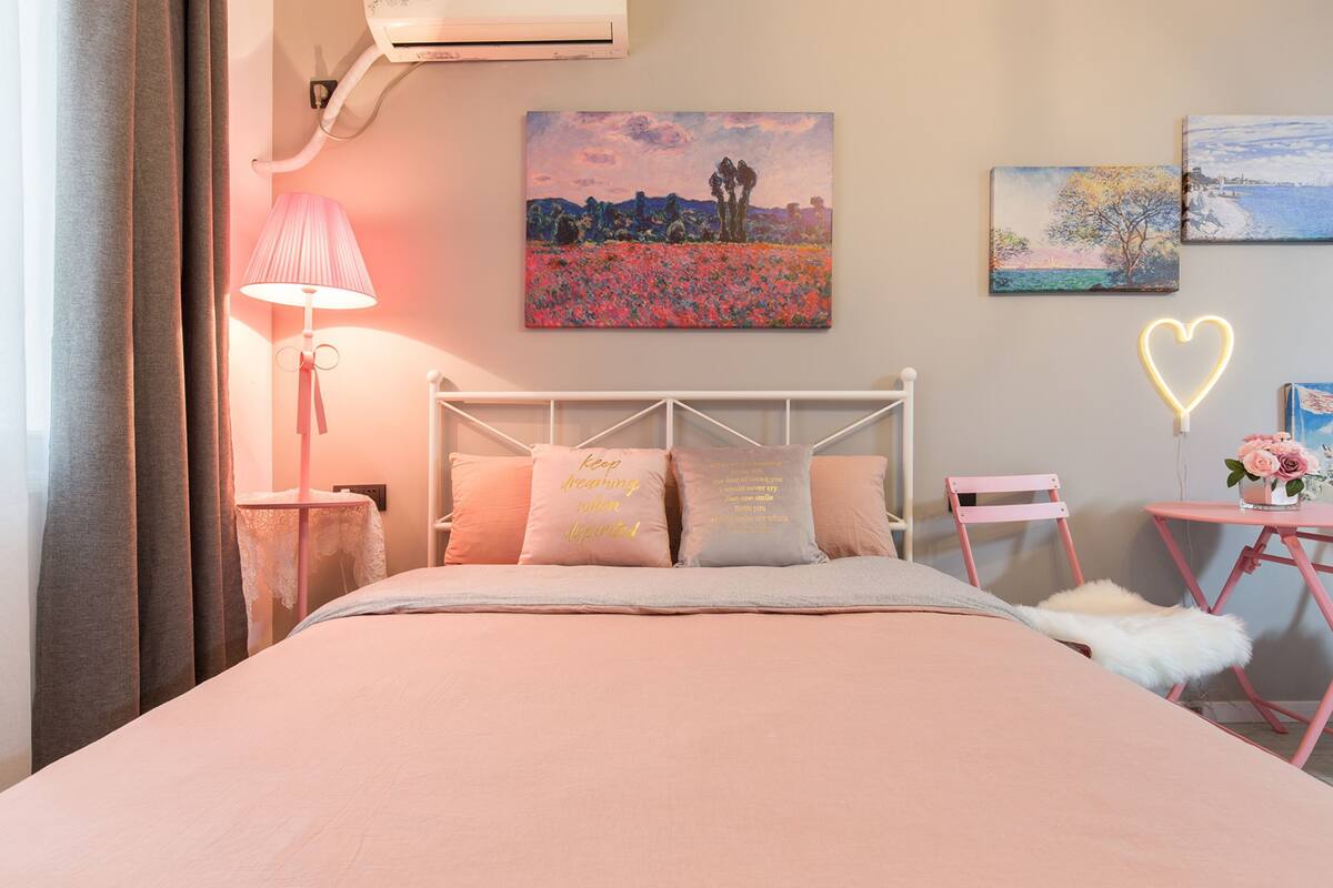 A bedroom with a bed, pillows, a bedside table, a lamp, a chair, a small table with flowers, and two paintings on the wall.