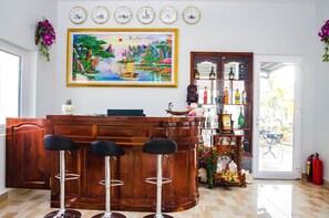 A bar area with a wooden counter, three black bar stools, a large framed painting, and a shelf with bottles and flowers.